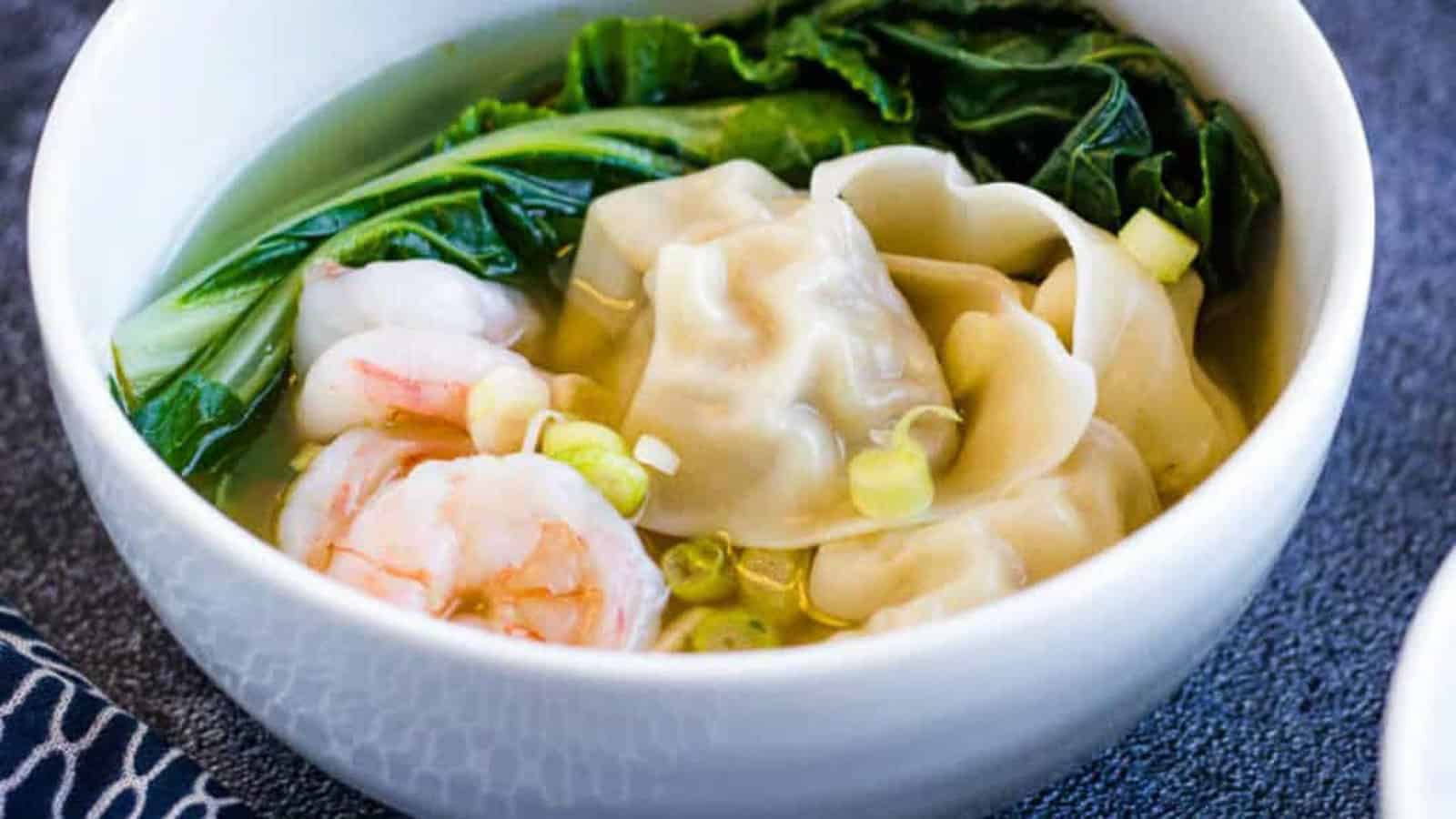 A bowl of wonton soup with dumplings, shrimp, leafy greens, and chopped green onions in clear broth.