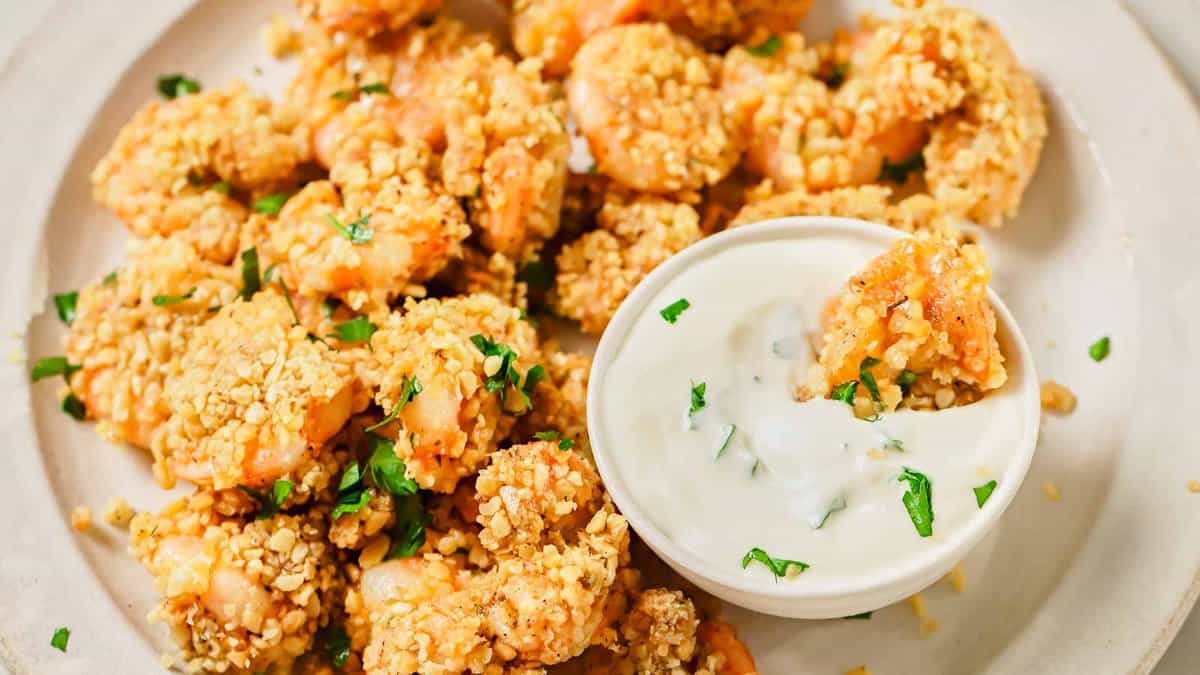 Plate of breaded shrimp garnished with chopped herbs, served with a small bowl of creamy dipping sauce.