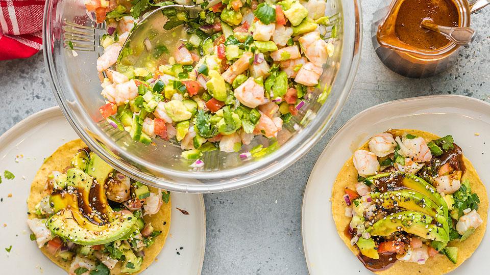 Two shrimp and avocado tostadas on plates beside a bowl of shrimp ceviche and a jar of brown sauce on a gray surface.