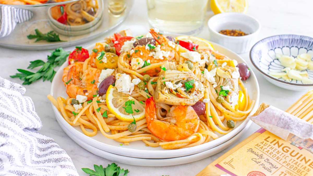 A plate of linguine pasta topped with shrimp, artichokes, feta cheese, lemon slices, olives, and parsley, with a side of sliced almonds and a partially visible pasta package.