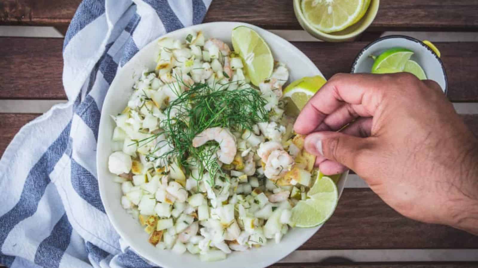 A hand adds a chip to a bowl of shrimp ceviche garnished with fresh dill and lime wedges, with a striped towel and lemon slices beside the bowl.