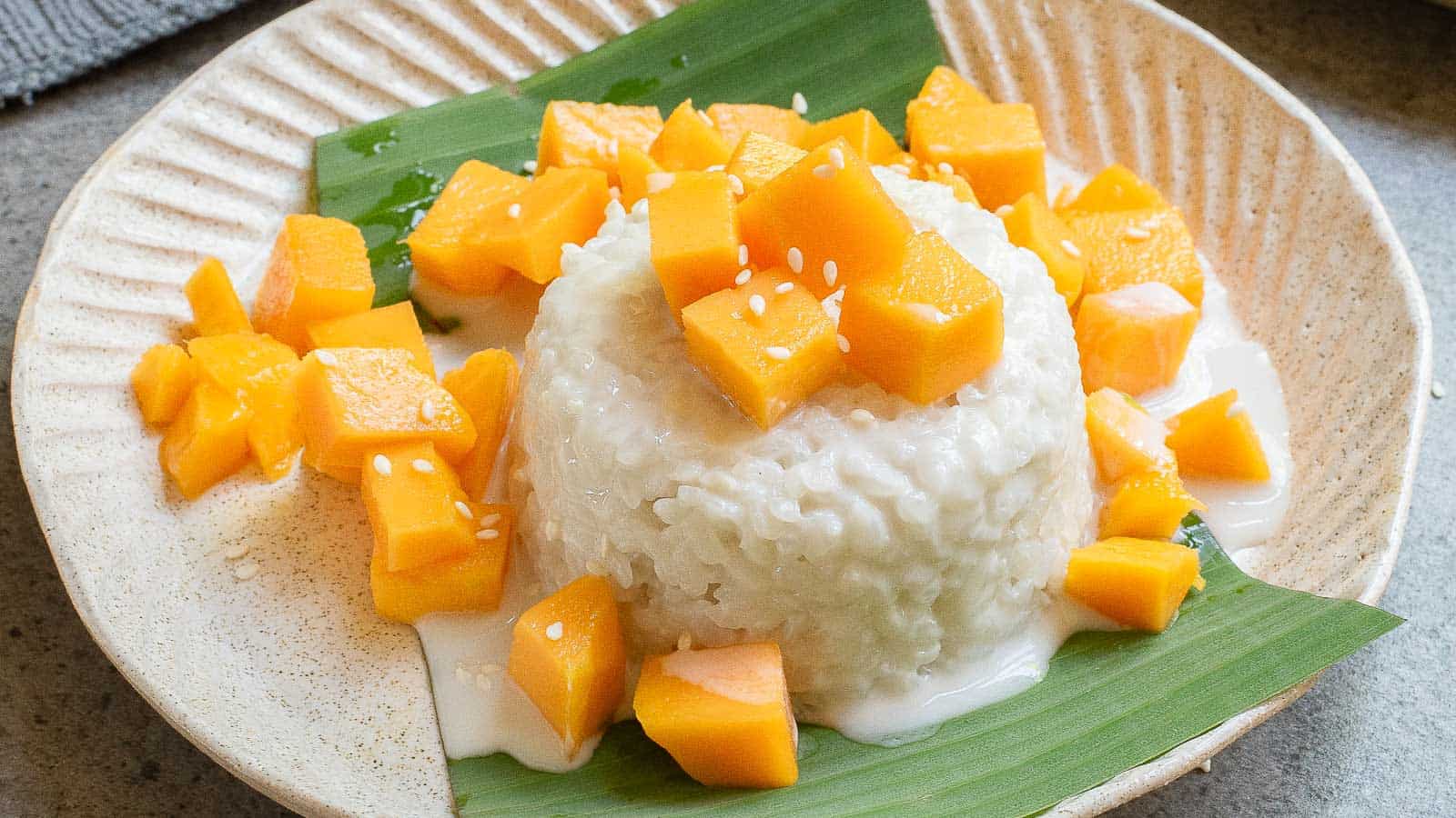 A plate of mango sticky rice with coconut sauce, topped with diced mango and sesame seeds, served on a green leaf.