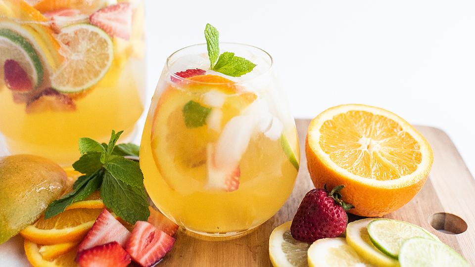 A glass of fruit-infused drink with ice, garnished with mint, sits on a wooden board with sliced citrus fruits and strawberries. A pitcher of the same drink is in the background.