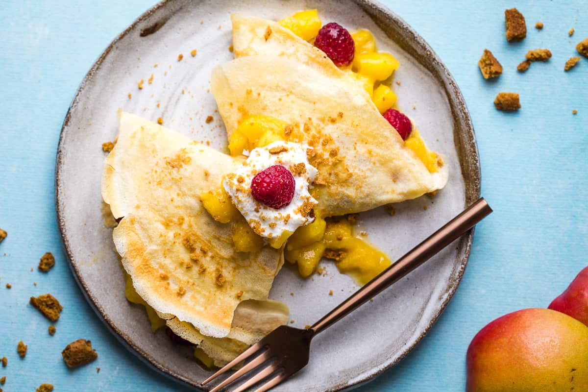 Two folded crepes filled with diced mango and topped with whipped cream, a raspberry, and crumbled cookies are served on a plate with a fork. Mango pieces and crumbs are scattered around.