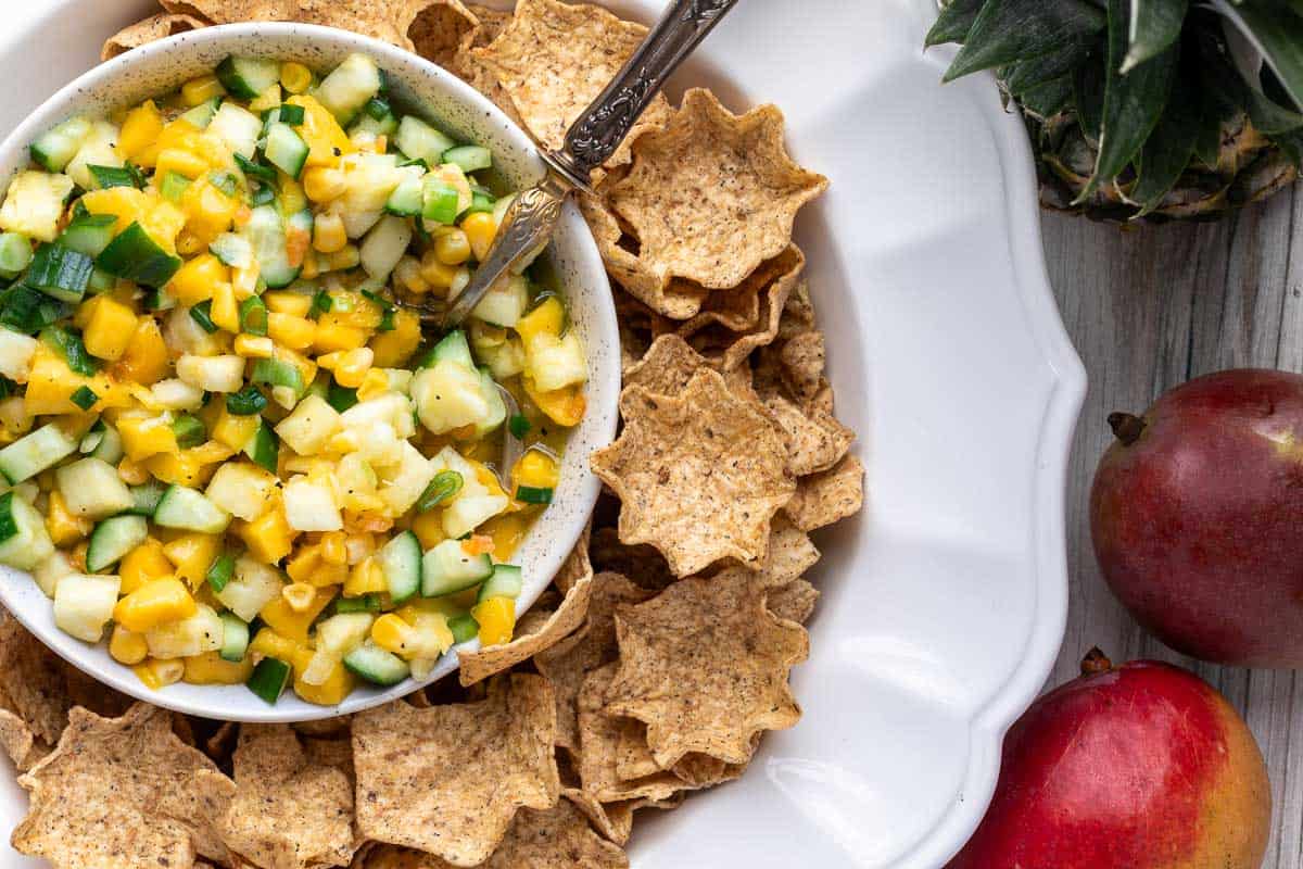 A bowl of pineapple mango salsa with diced cucumbers and green onions, surrounded by star-shaped tortilla chips on a white plate, with mangoes and a pineapple beside the plate.