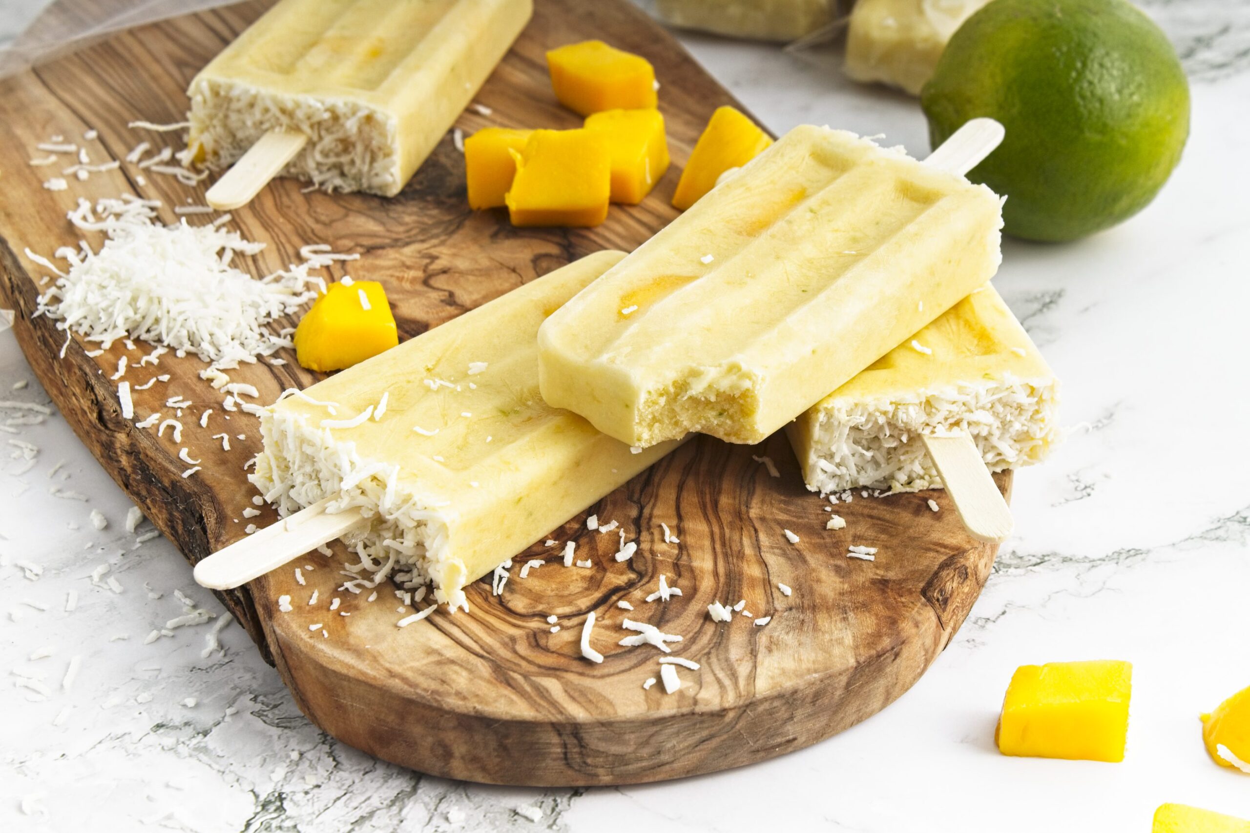 Three popsicles with shredded coconut on a wooden board, surrounded by mango cubes, shredded coconut, and a whole lime on a marble surface.