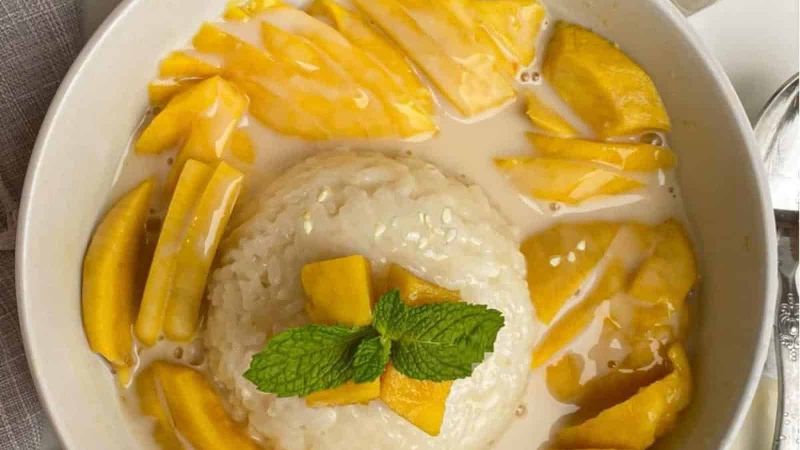 A bowl of mango sticky rice topped with coconut sauce, garnished with a sprig of mint and sesame seeds, surrounded by fresh mango slices.