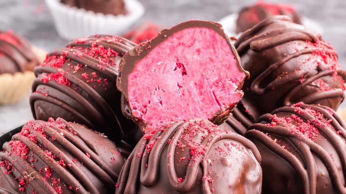 A close-up of chocolate truffles with a pink filling, drizzled with chocolate and sprinkled with red crumbs, one truffle cut open to show the inside.