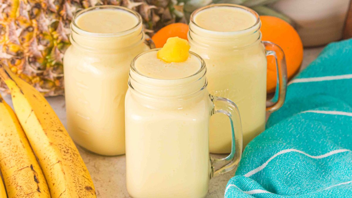 Three mason jar mugs filled with creamy yellow smoothies are placed on a counter, surrounded by bananas, a pineapple, oranges, and a turquoise towel.