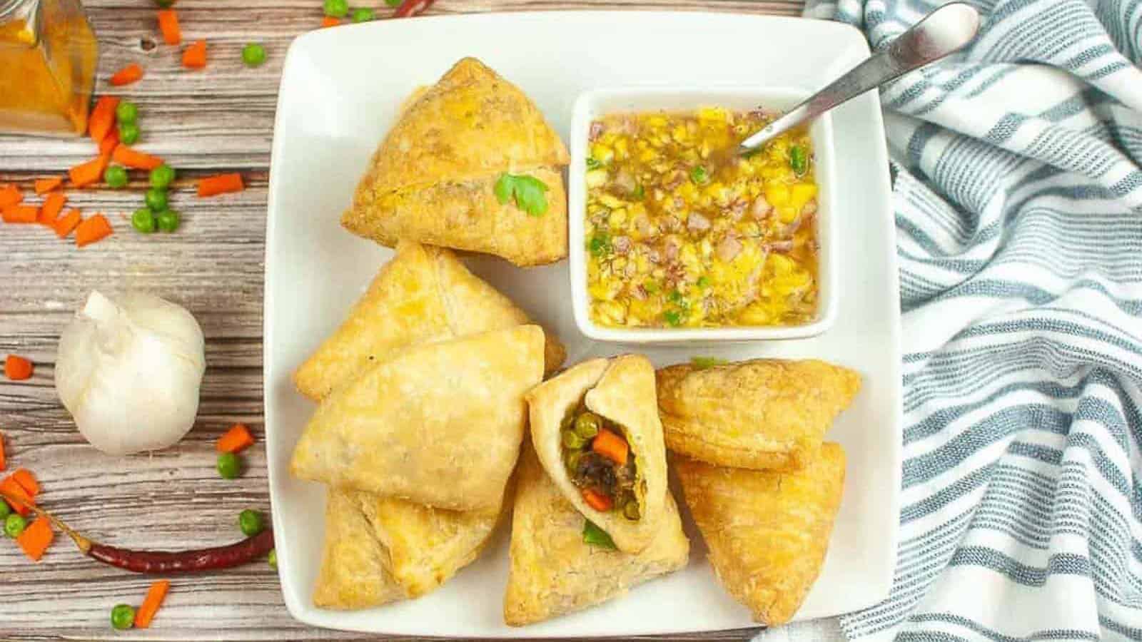A plate of golden-brown samosas with visible vegetable filling, served with a small bowl of chutney; scattered vegetables and a garlic bulb are nearby.