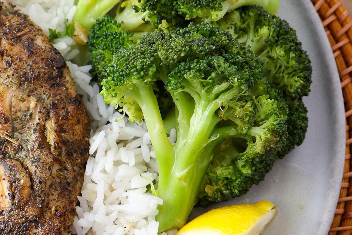 Close-up of a plate with cooked broccoli, white rice, a slice of seasoned meat, and a lemon wedge.