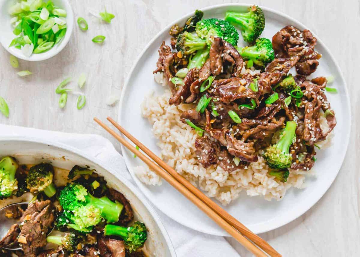 A plate of beef and broccoli stir-fry served over white rice, garnished with green onions, with chopsticks placed on the side.