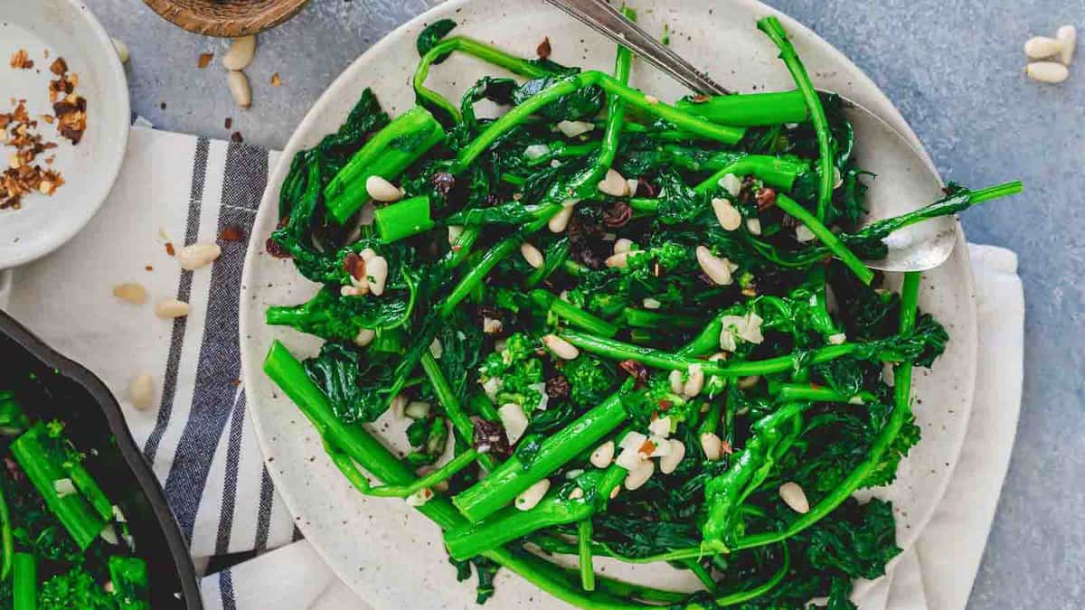 A plate of sautรฉed broccoli rabe garnished with pine nuts and raisins, served with a spoon on a striped napkin.