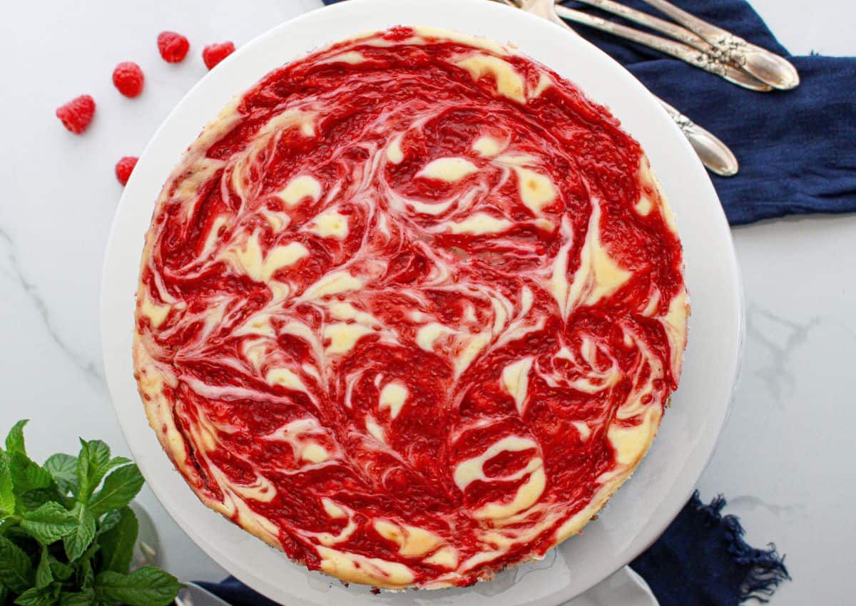 A raspberry swirl cheesecake sits on a white plate with a marbled red and white pattern on top, surrounded by fresh raspberries, mint, and silver utensils.