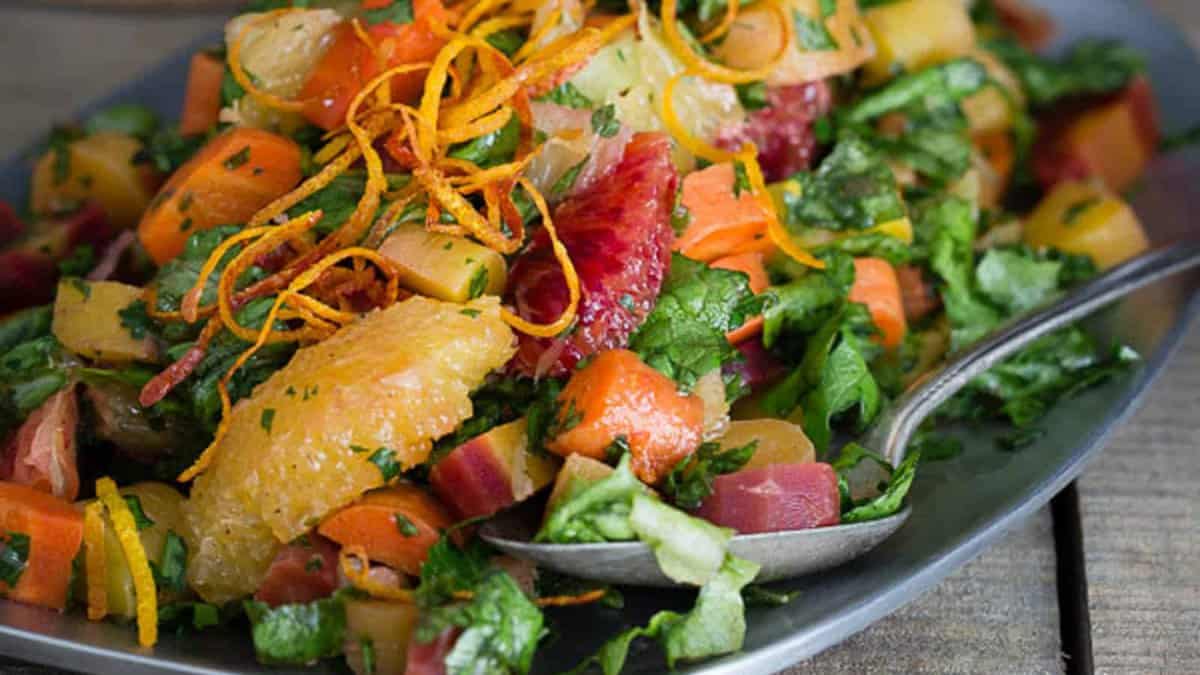 A platter of mixed salad with citrus segments, chopped carrots, leafy greens, and thin orange peel strips, served with a spoon.