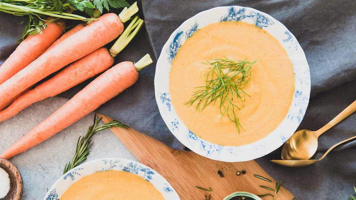 A bowl of creamy orange soup garnished with fresh herbs sits next to whole carrots, a spoon, and a napkin on a wooden surface.