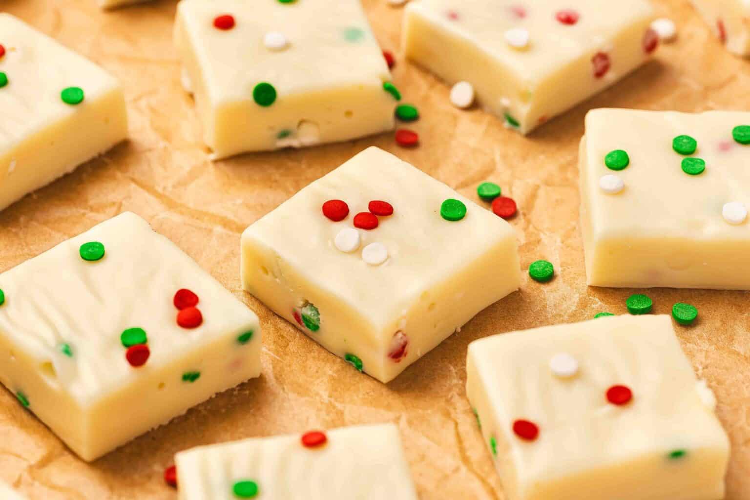 Squares of white fudge topped with red, green, and white sprinkles are arranged on brown parchment paper.