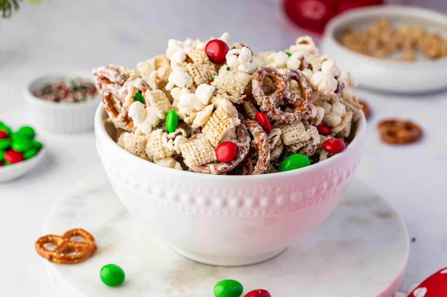 A white bowl filled with a snack mix of Chex cereal, pretzels, popcorn, and red and green candy-coated chocolates on a white surface.