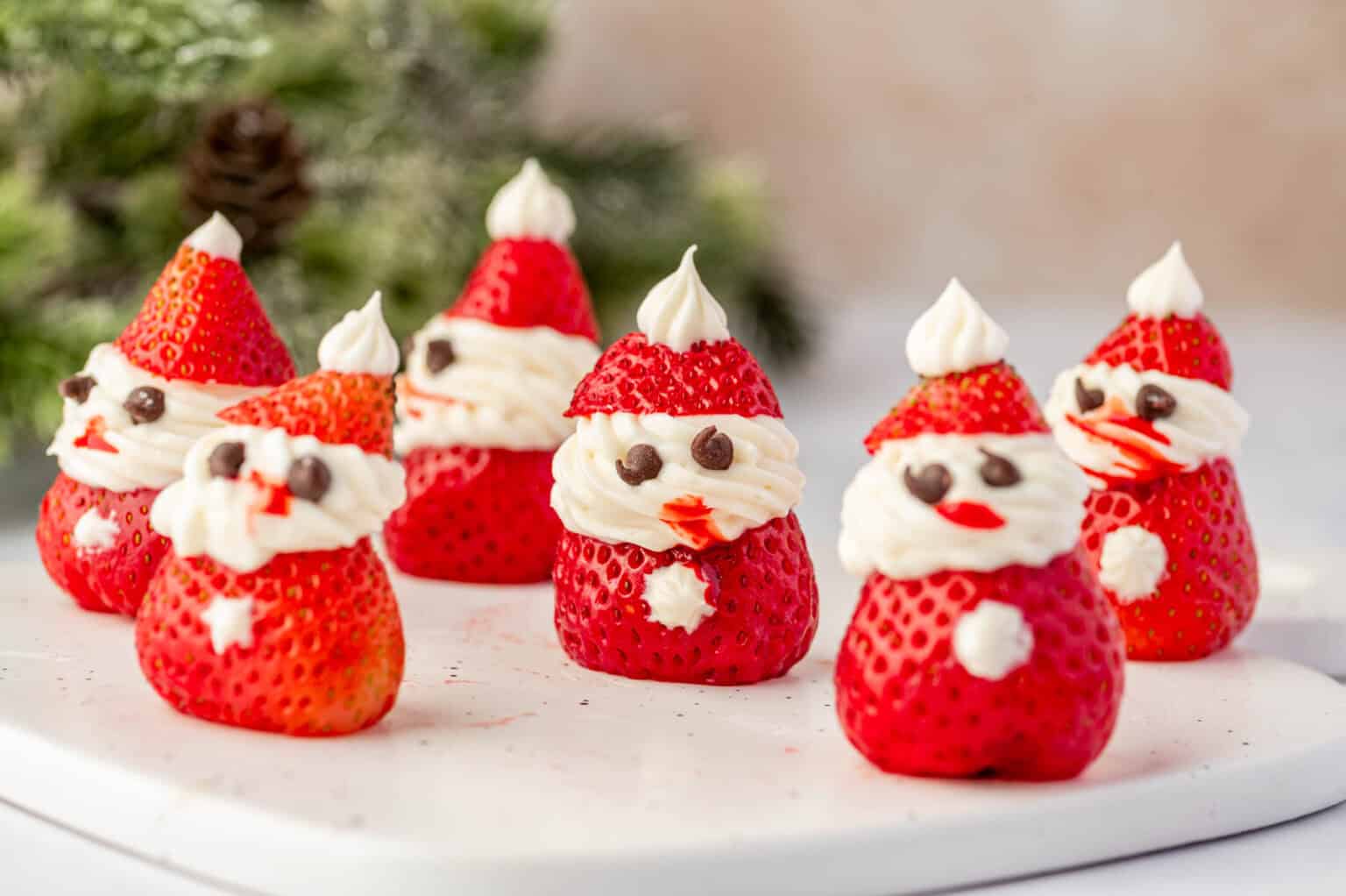 Six strawberries decorated with whipped cream and chocolate chips to look like Santa Claus, arranged on a white surface with greenery in the background.