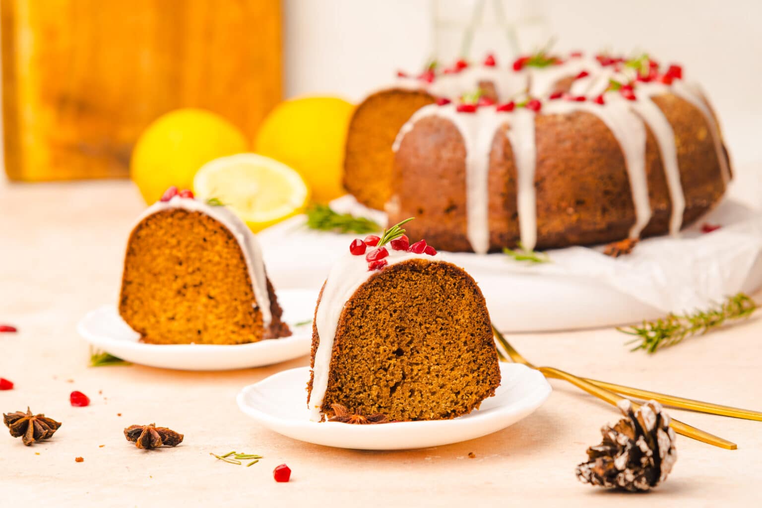 A bundt cake with white icing and pomegranate seeds sits on a table, with two slices served on small plates. Lemons, herbs, and decorative items are in the background.