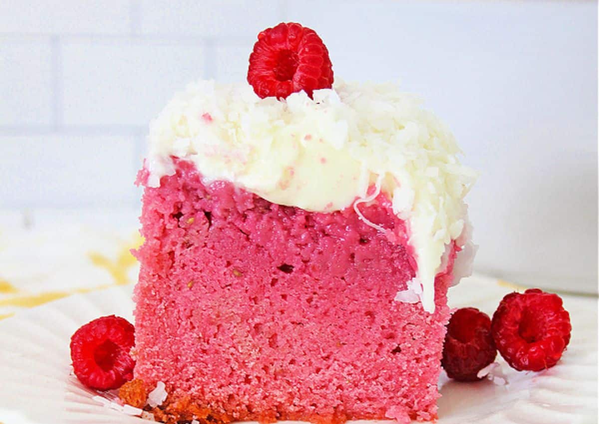 A slice of pink cake topped with white frosting, shredded coconut, and a raspberry, garnished with additional raspberries on a white plate.