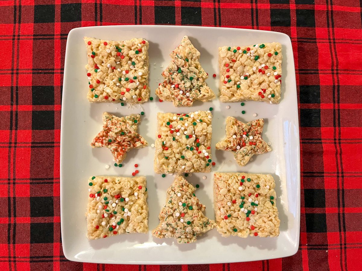 A square white plate with nine Rice Krispies treats, some cut into stars and trees, all topped with red, green, and white sprinkles, on a red and black plaid tablecloth.