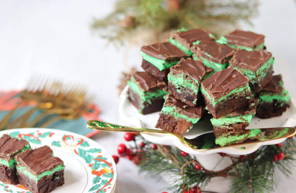 A plate of stacked chocolate mint brownies with green mint filling sits on a white cake stand, surrounded by festive holiday decorations and a plate with forks.