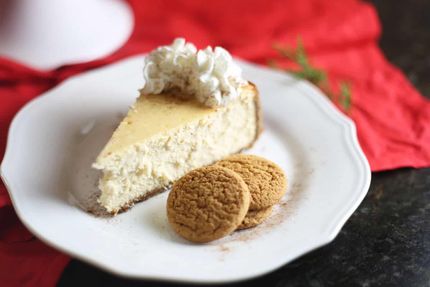A slice of cheesecake topped with whipped cream sits on a white plate next to two round cookies, with a red cloth in the background.