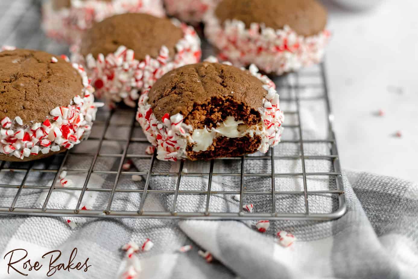 Chocolate cookies filled with white cream and rolled in crushed peppermint candy, displayed on a cooling rack; one cookie has a bite taken out of it.