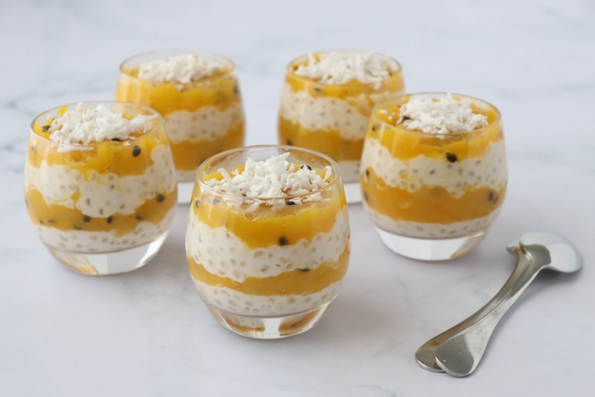 Five glass cups with layered mango and tapioca pudding, topped with shredded coconut, are arranged on a white surface next to two metal dessert spoons.