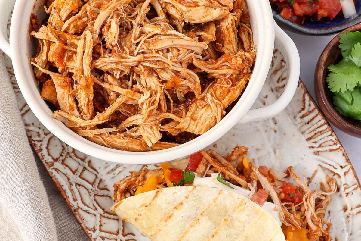 A bowl of shredded chicken sits on a plate next to a taco filled with shredded chicken, diced vegetables, and cheese. Fresh cilantro and salsa are visible on the side.