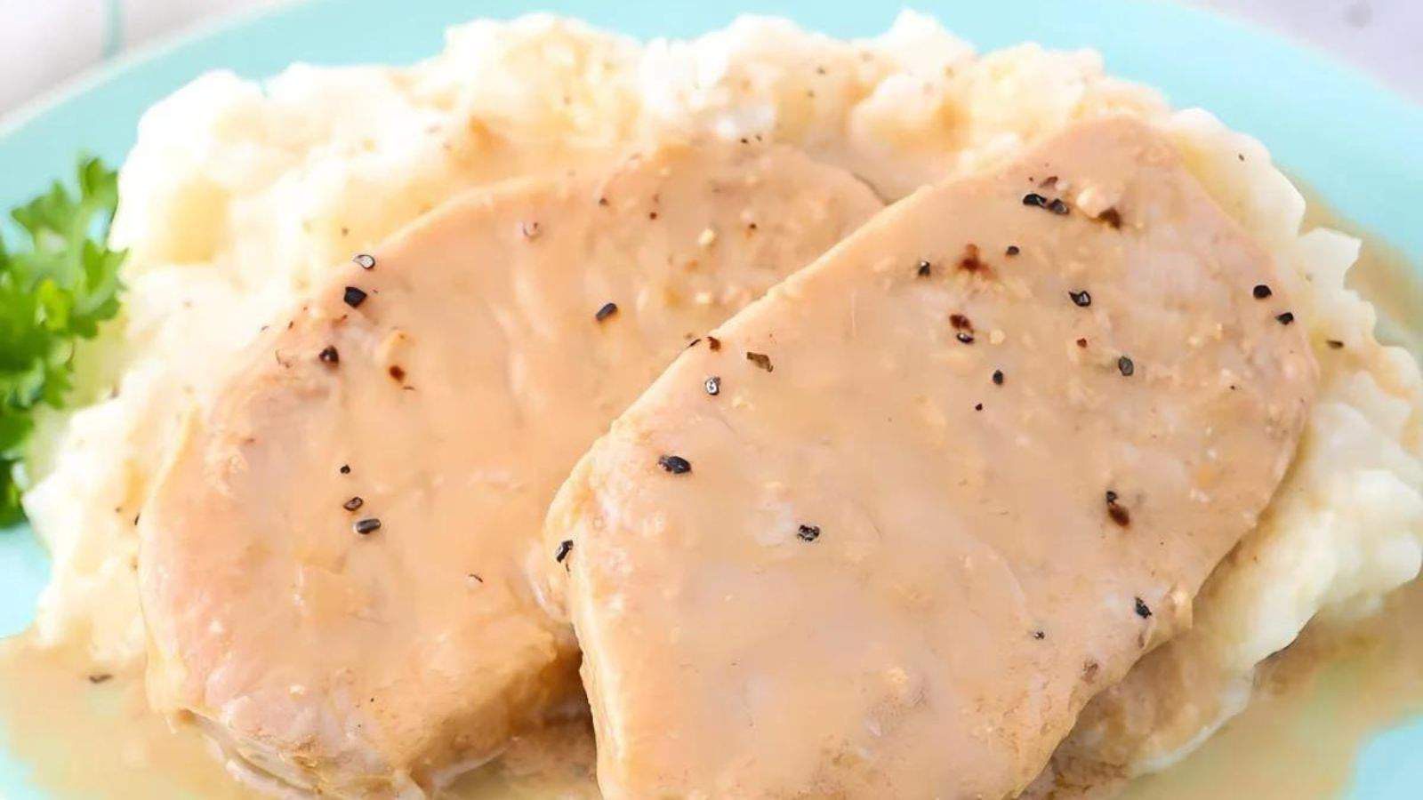 Two slices of pork covered in gravy are served on a bed of mashed potatoes, garnished with a sprig of parsley.