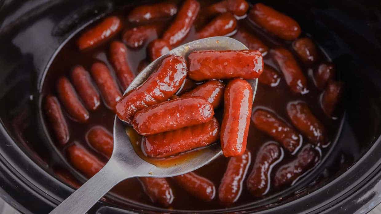 A spoon holds several cocktail sausages coated in sauce above a slow cooker filled with more sausages in sauce.