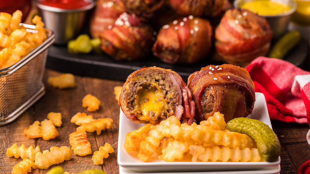 A plate with crinkle-cut fries, bacon-wrapped meatballs filled with cheese, and a pickle slice. More fries and sauces are visible in the background.