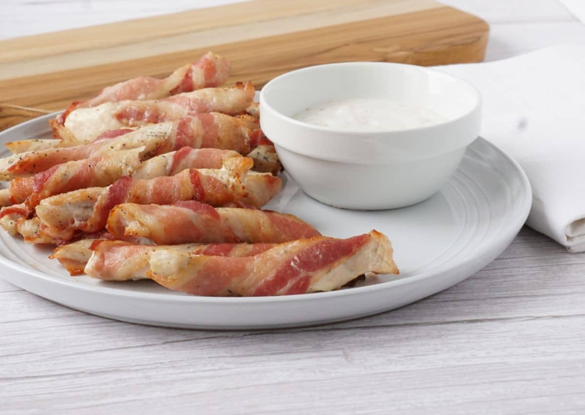 A white plate with bacon-wrapped chicken strips and a small bowl of white dipping sauce, set on a light wooden table.