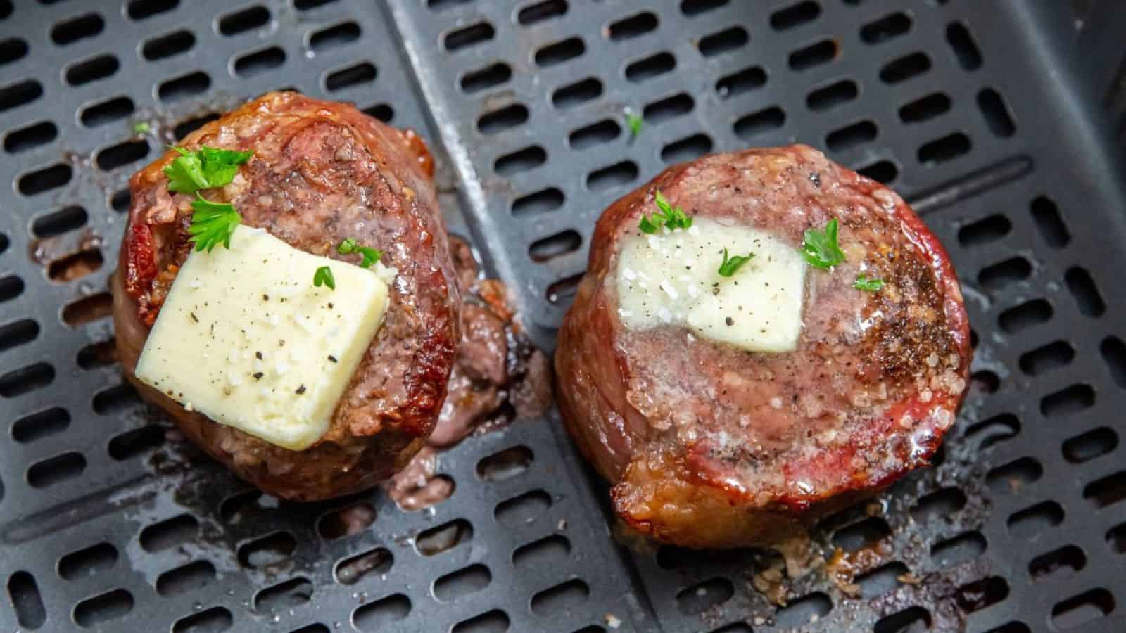 Two cooked filet mignon steaks in an air fryer basket, each topped with a pat of melting butter and garnished with chopped parsley.