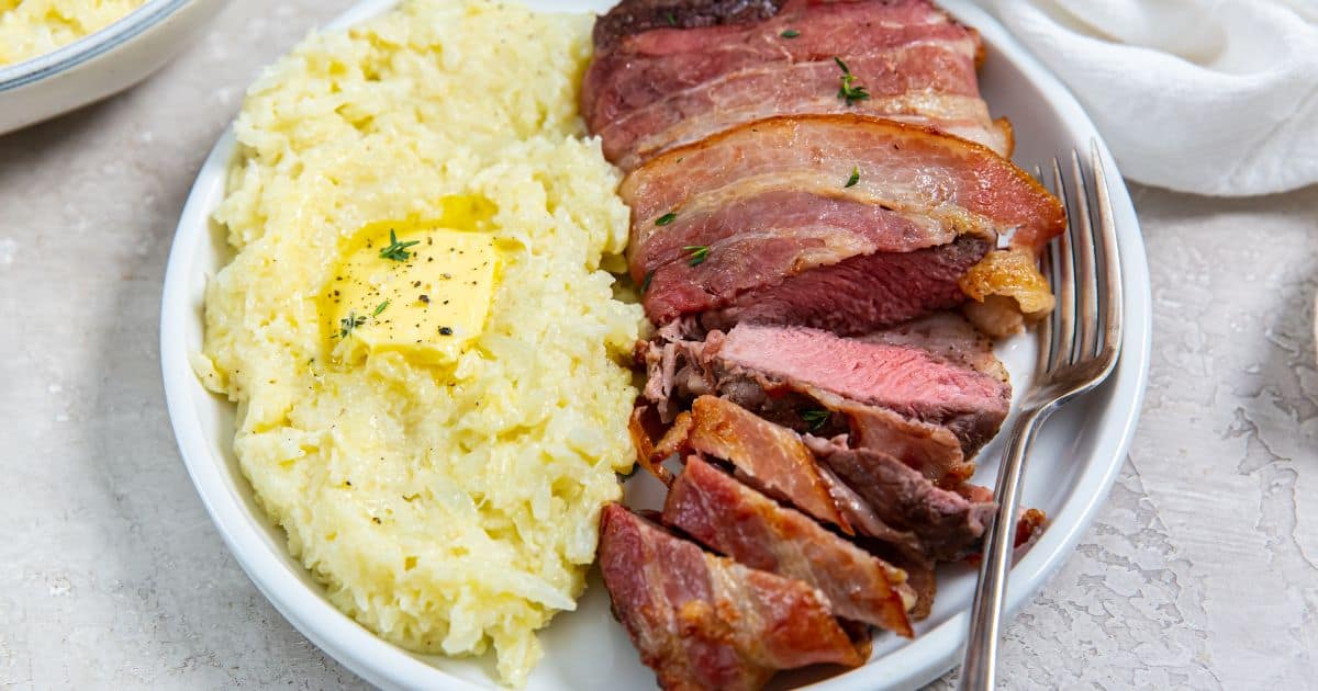 A plate with sliced bacon-wrapped steak and a serving of mashed potatoes topped with butter and black pepper, with a fork on the side.