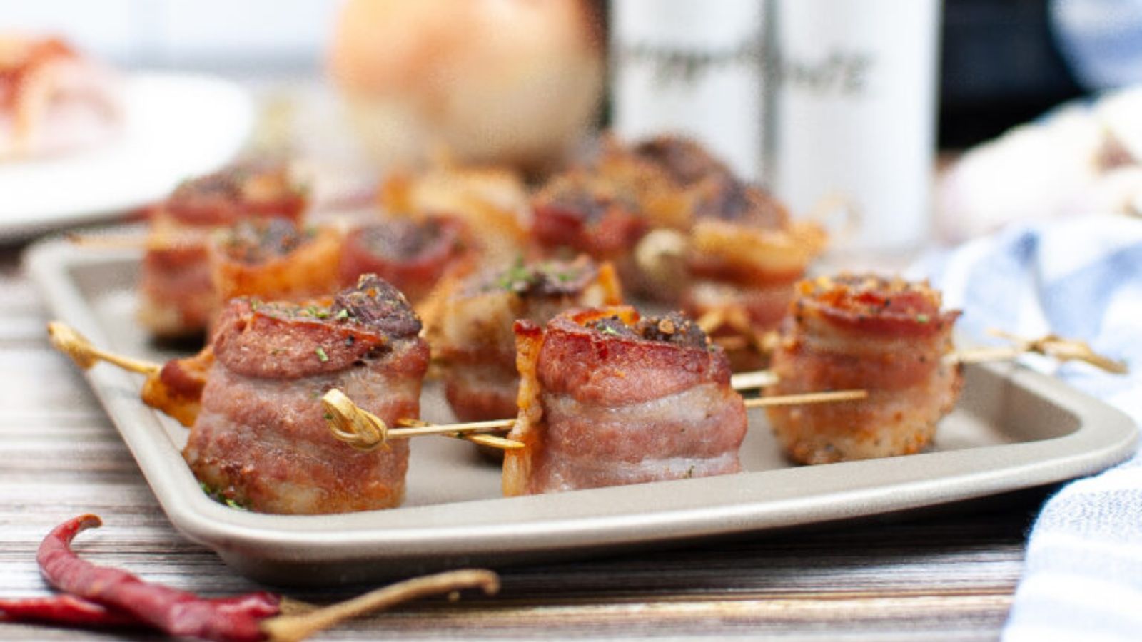A baking tray with bacon-wrapped appetizers secured with toothpicks, garnished with herbs, sits on a wooden surface.