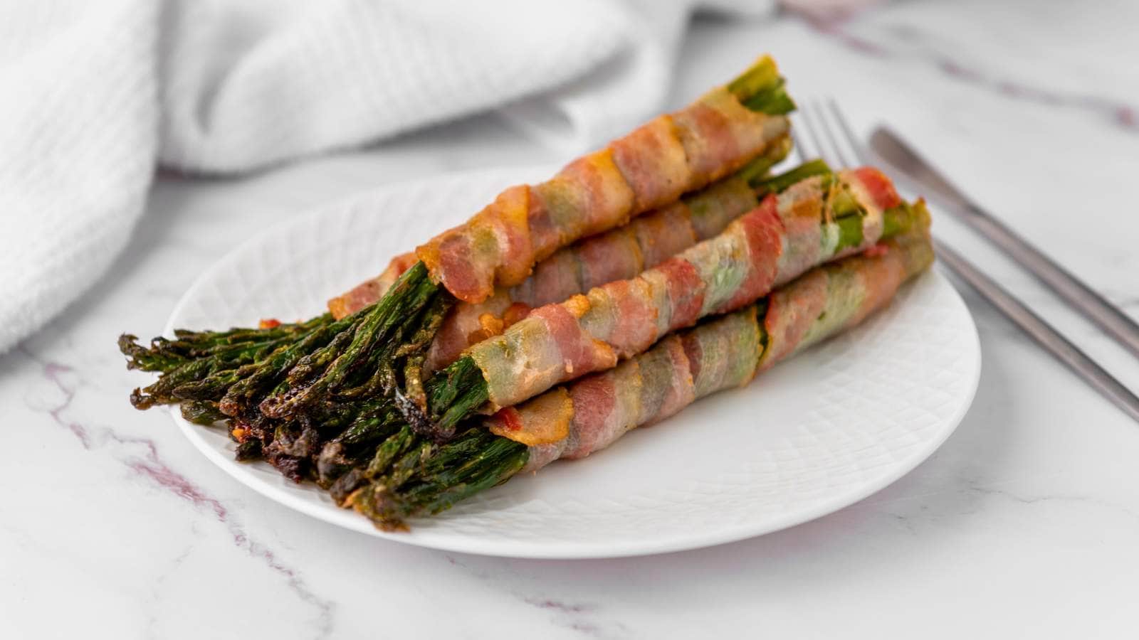Four bundles of asparagus spears wrapped in cooked bacon are arranged on a white plate, with a fork and knife placed beside the plate.