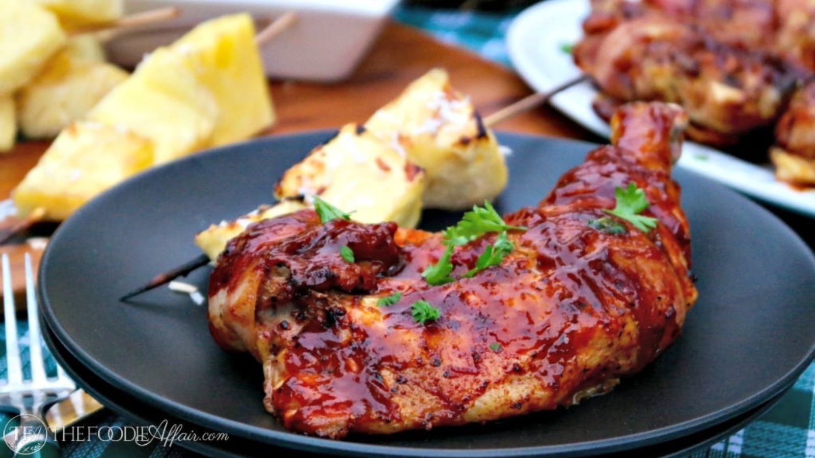 Grilled chicken leg coated in barbecue sauce, garnished with parsley, served with skewered grilled pineapple on a black plate.