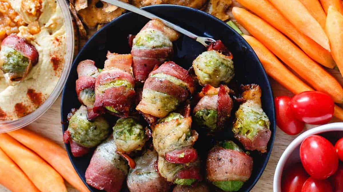 A bowl of bacon-wrapped Brussels sprouts surrounded by carrots, grape tomatoes, and a dish of hummus with crackers.