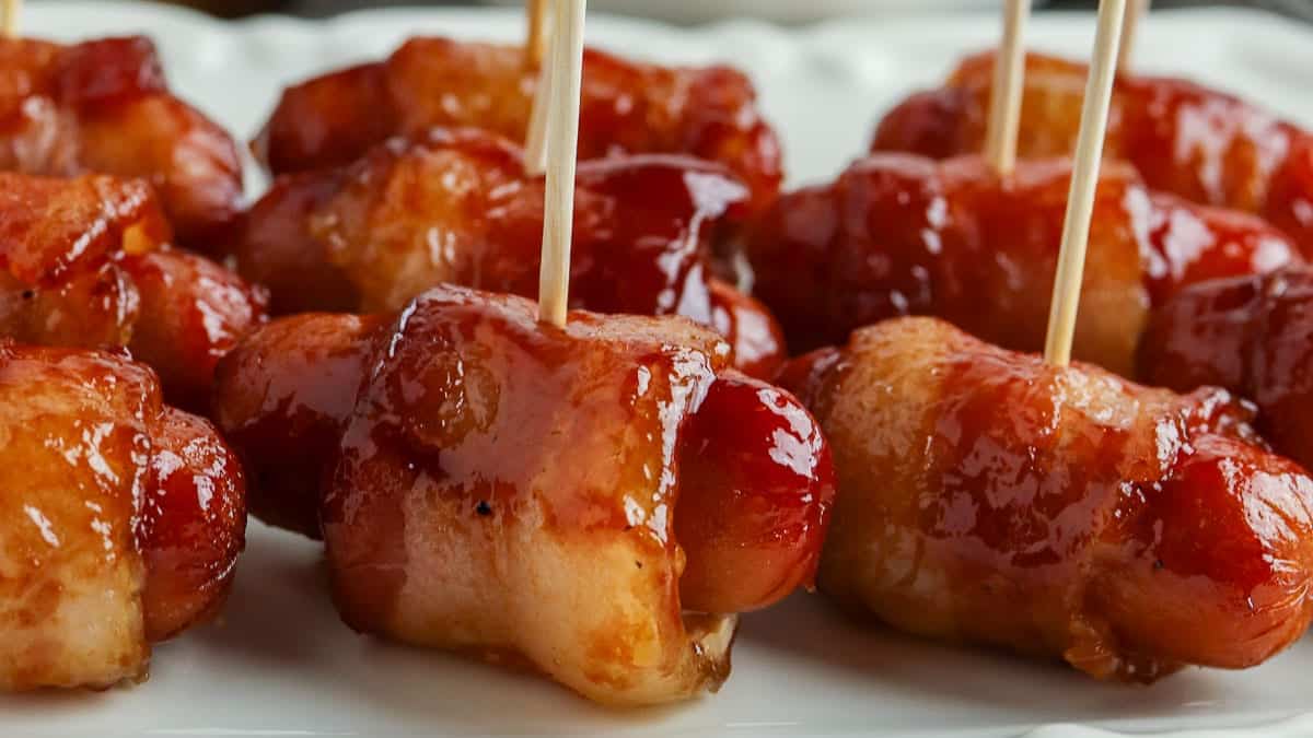 Close-up of bacon-wrapped cocktail sausages secured with toothpicks, arranged on a white plate.