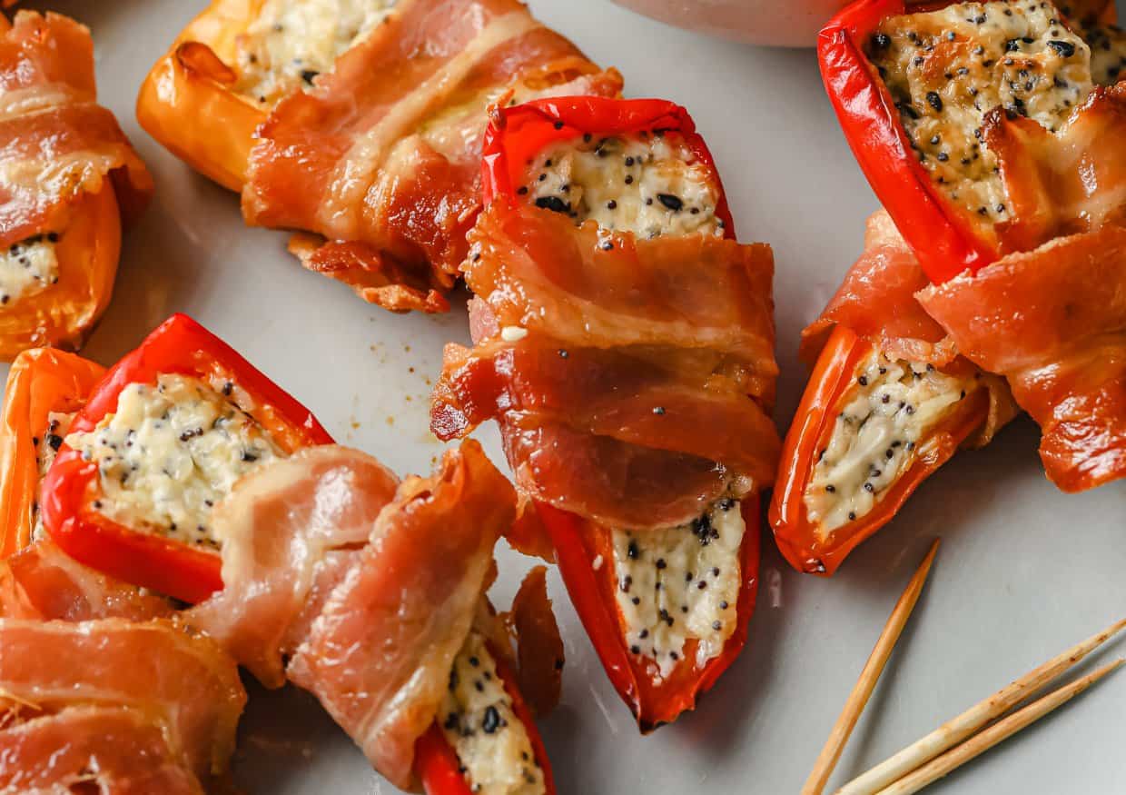 Mini red peppers stuffed with a creamy cheese mixture and wrapped in bacon, arranged on a plate with toothpicks nearby.