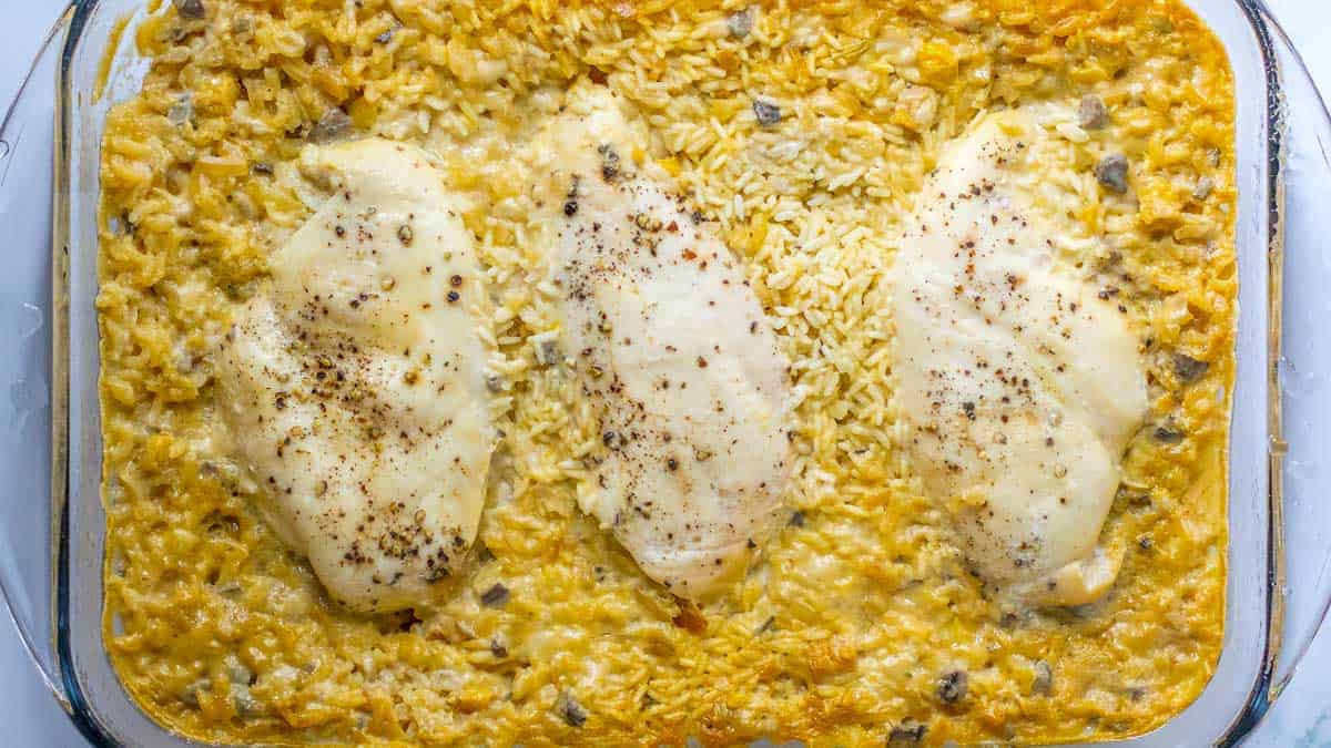 A glass baking dish filled with baked chicken breasts atop a creamy mixture of rice and sauce, seasoned with pepper.