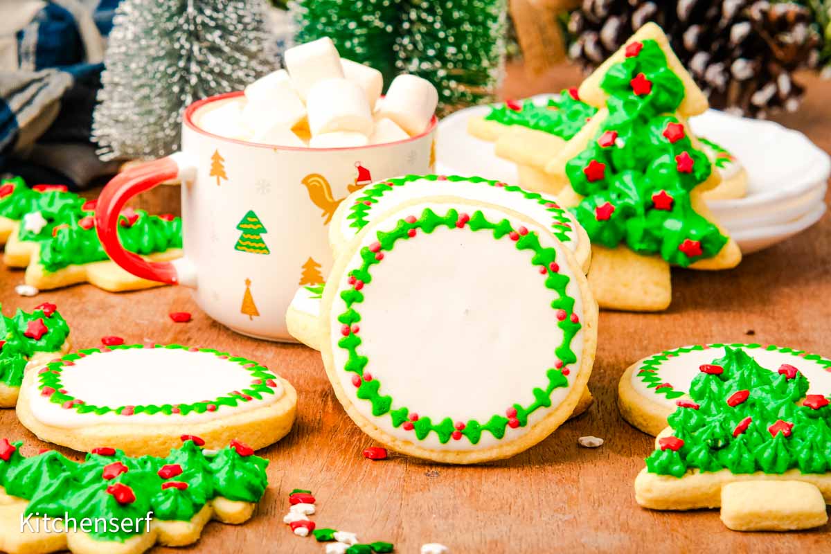 Decorated sugar cookies with holiday designs and a mug of hot chocolate with marshmallows on a wooden table, with a festive background.