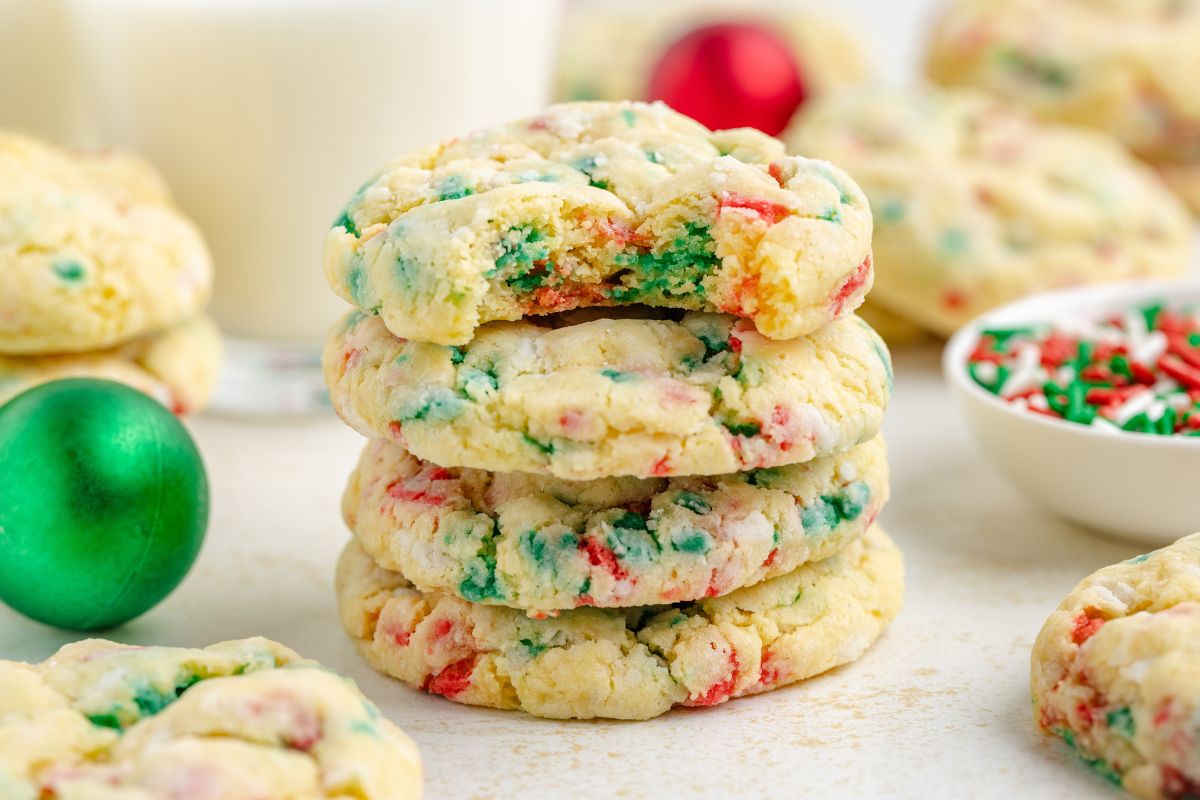A stack of three colorful sugar cookies with red and green sprinkles sits on a table, with a bitten cookie on top and holiday decorations in the background.