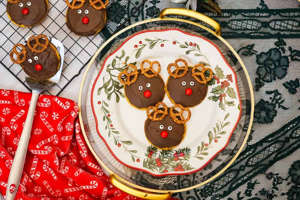 Chocolate-covered cookies decorated as reindeer with pretzel antlers, candy eyes, and red noses arranged on a festive plate and a cooling rack.