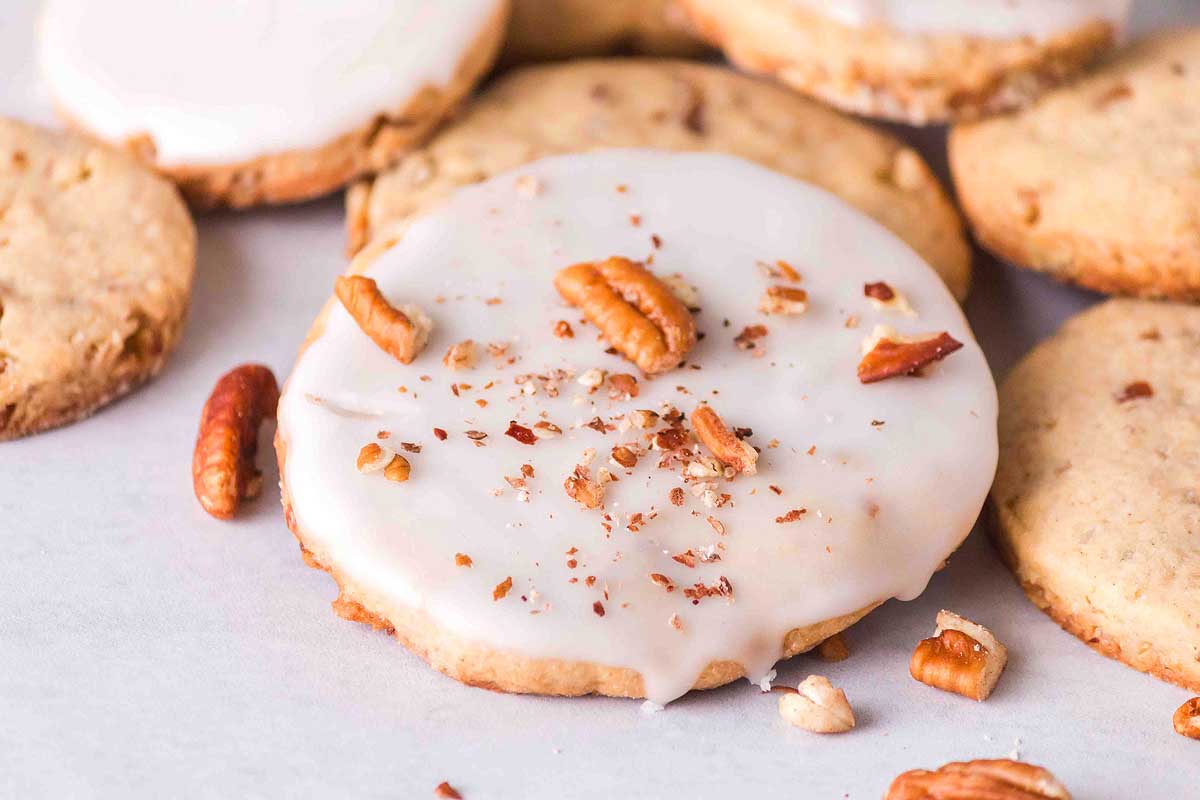 Round cookie with white icing and crushed pecans on top, surrounded by similar cookies on a light surface.