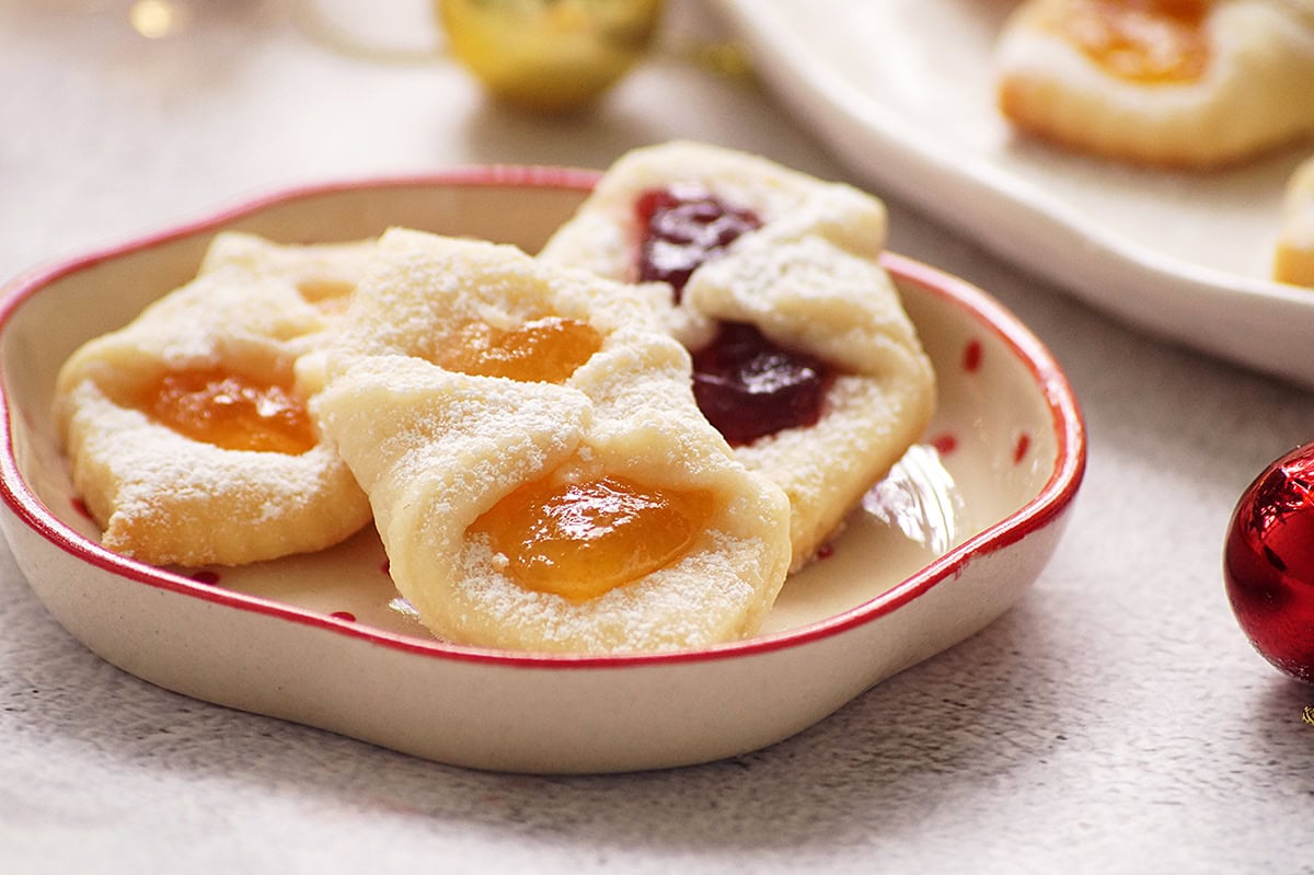 A plate of four pastries filled with yellow and red fruit jams, dusted with powdered sugar, on a light surface.