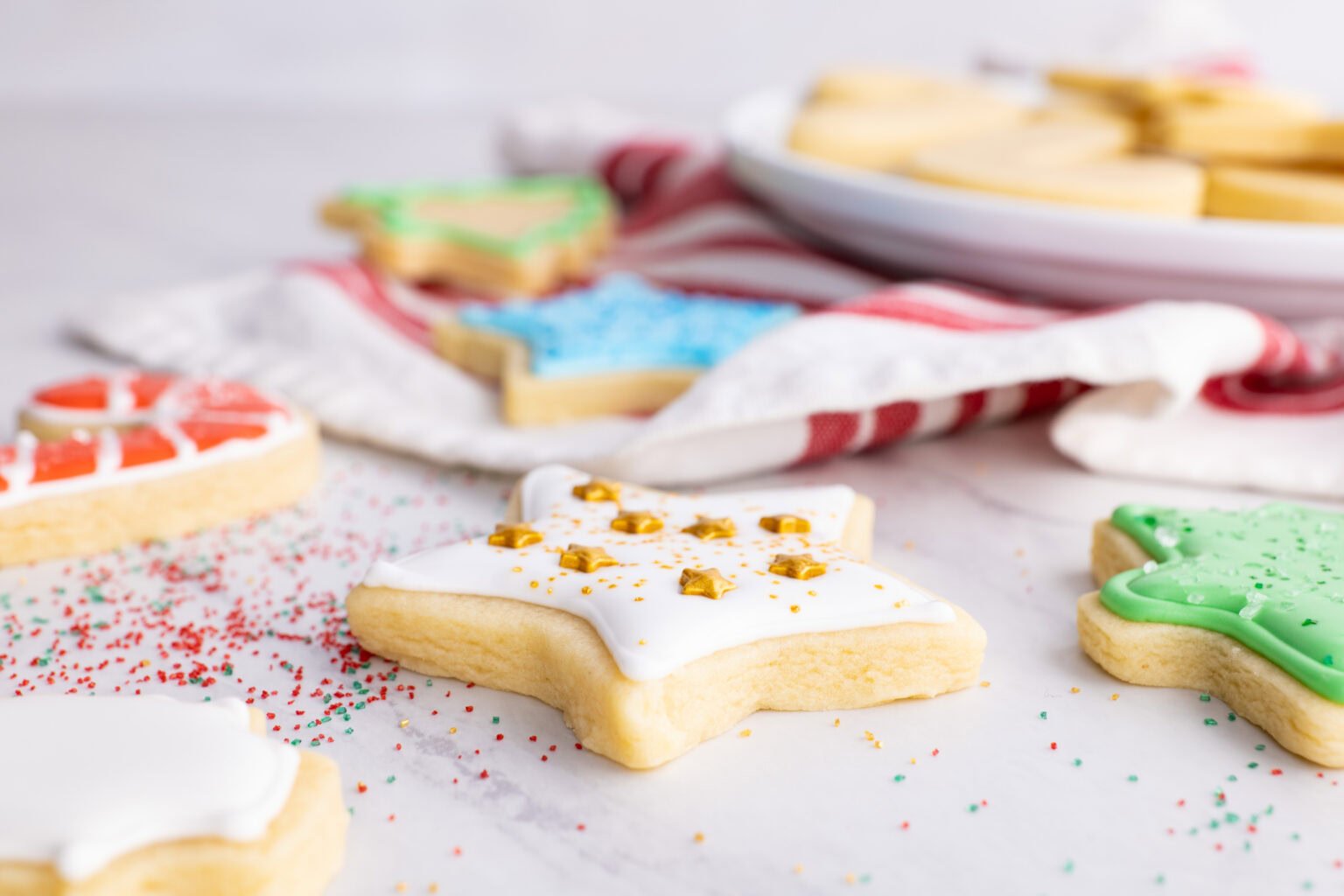 Decorated star-shaped sugar cookies with white icing and sprinkles on a marble surface, with more cookies and a red-striped cloth in the background.
